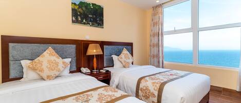 A hotel room with two beds, a painting on the wall, a nightstand with a lamp, and a large window with a view of the sea.