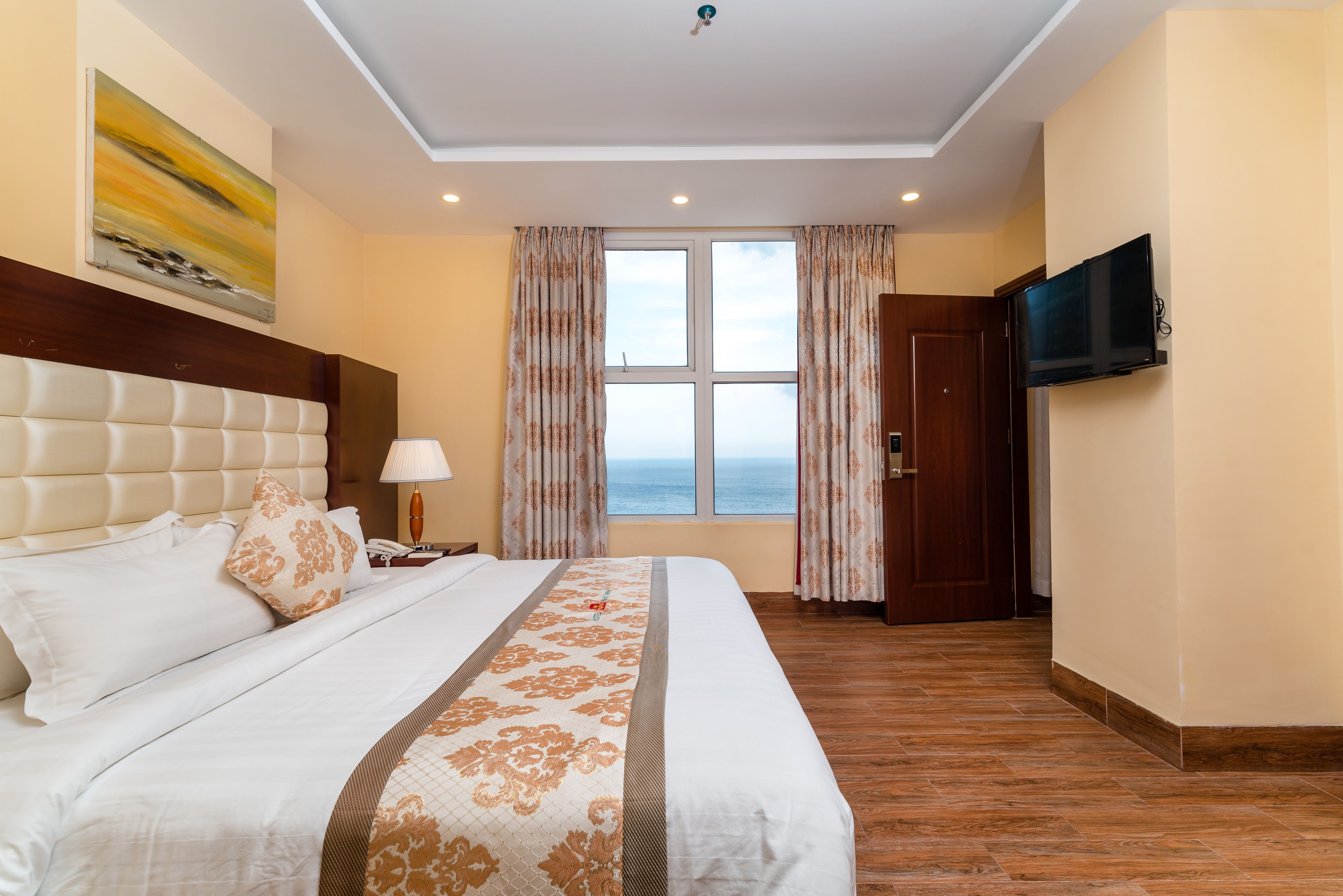 A hotel room with a large bed, a TV mounted on the wall, and a view of the sea through the window.
