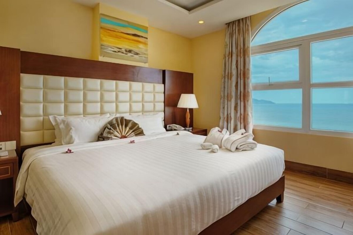 Habitación de hotel con una cama grande, cabecera, mesita de noche, lámpara y vistas al mar.