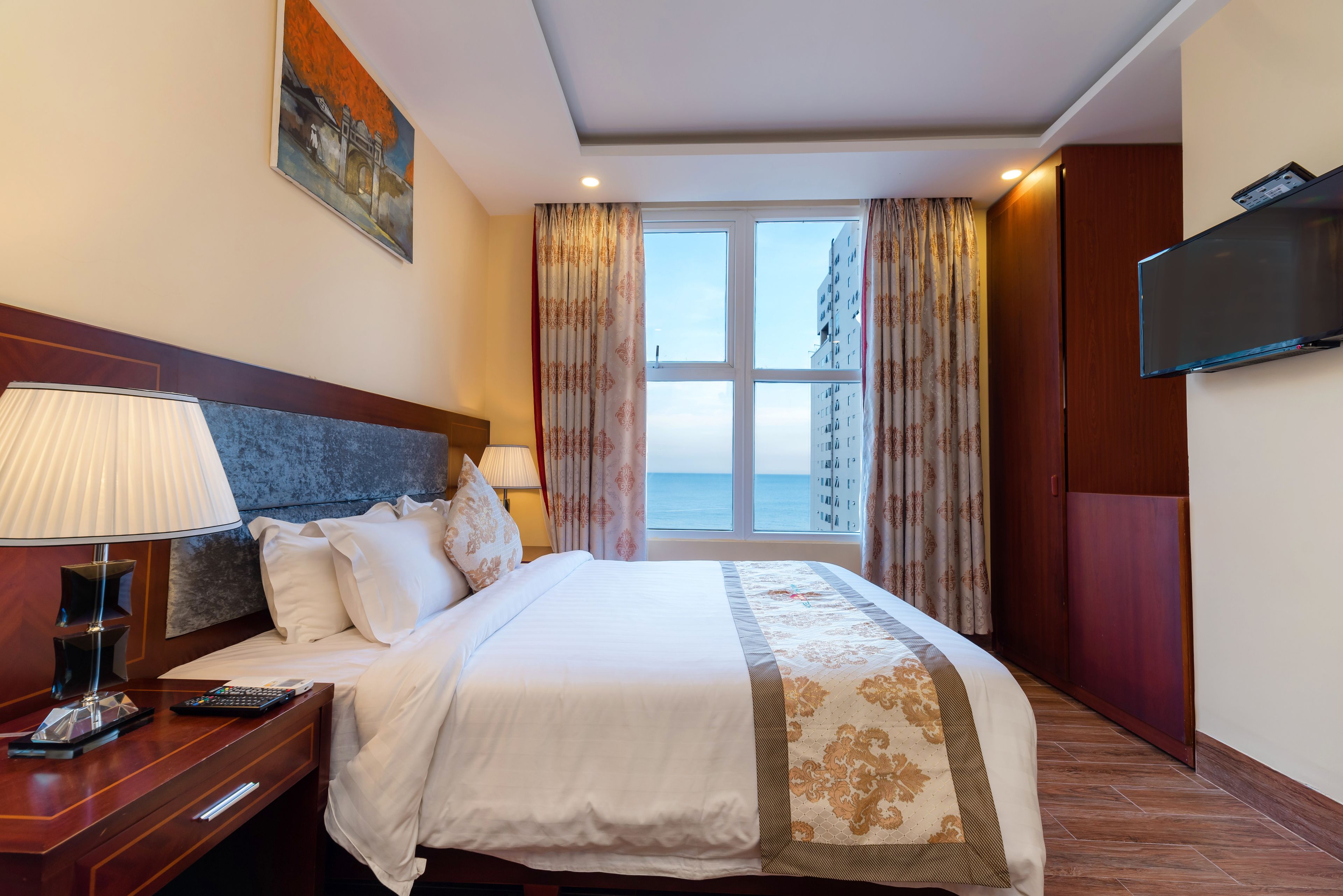 Habitación de hotel con una cama grande, un televisor montado en la pared, una mesita de noche con lámpara y una ventana con cortinas.