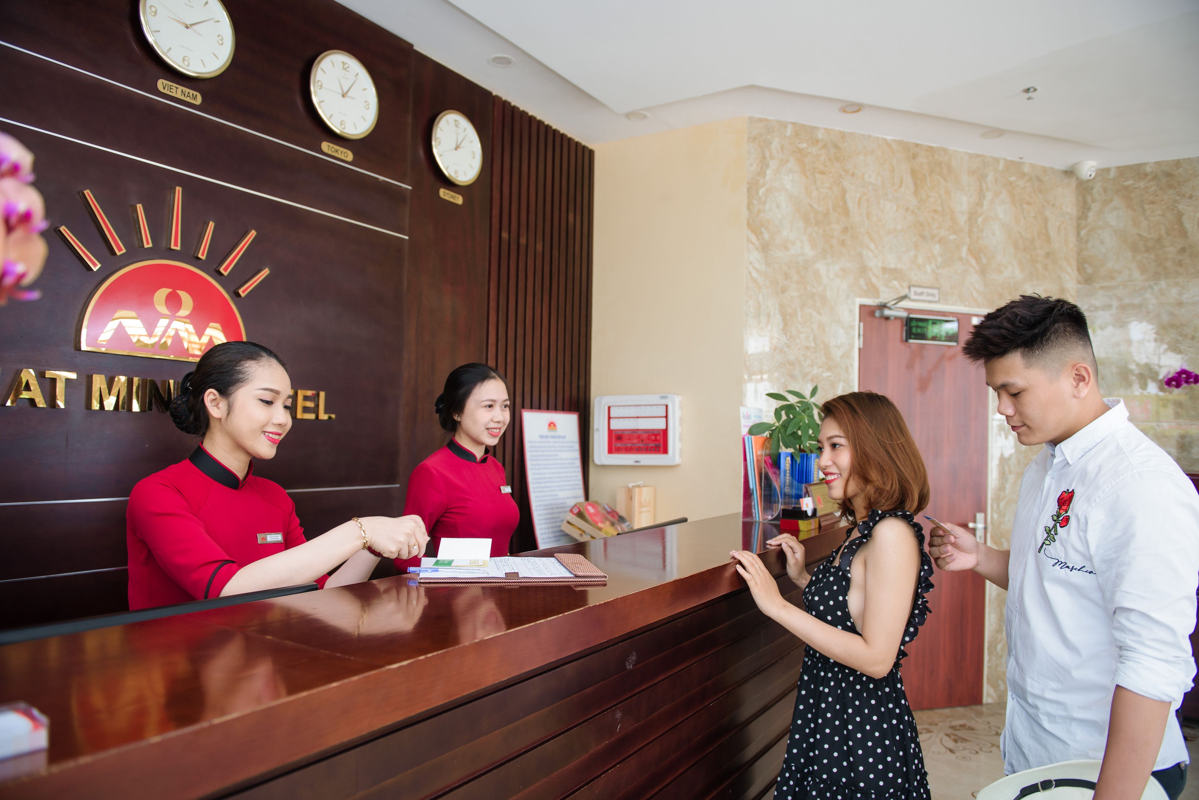Una mesa de recepción con un reloj, un cartel con la leyenda 'Viet Nam' y 'Tokyo', y una recepcionista con uniforme rojo.