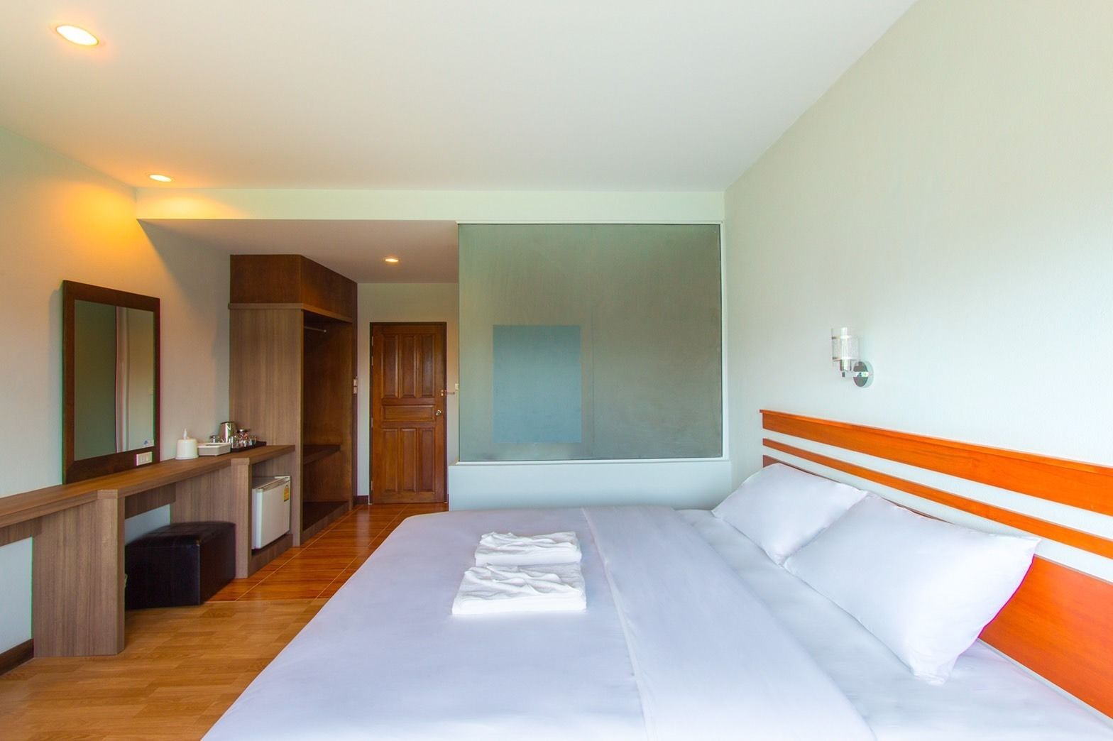 A hotel room with a large bed, a wooden headboard, a mirror, a desk with a chair, and a wardrobe.