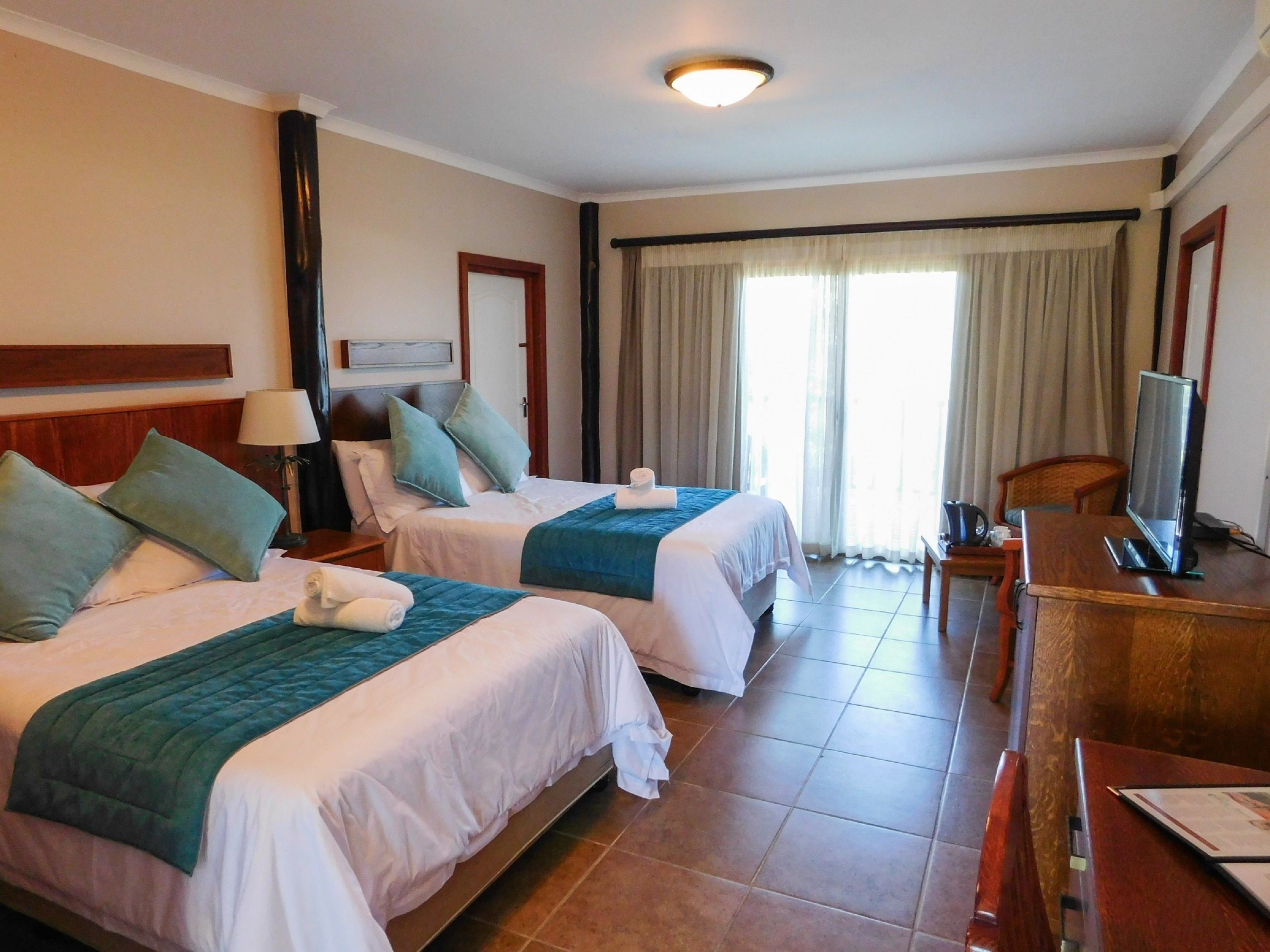 A hotel room with two beds, a desk with a computer, a TV, and a window with curtains.