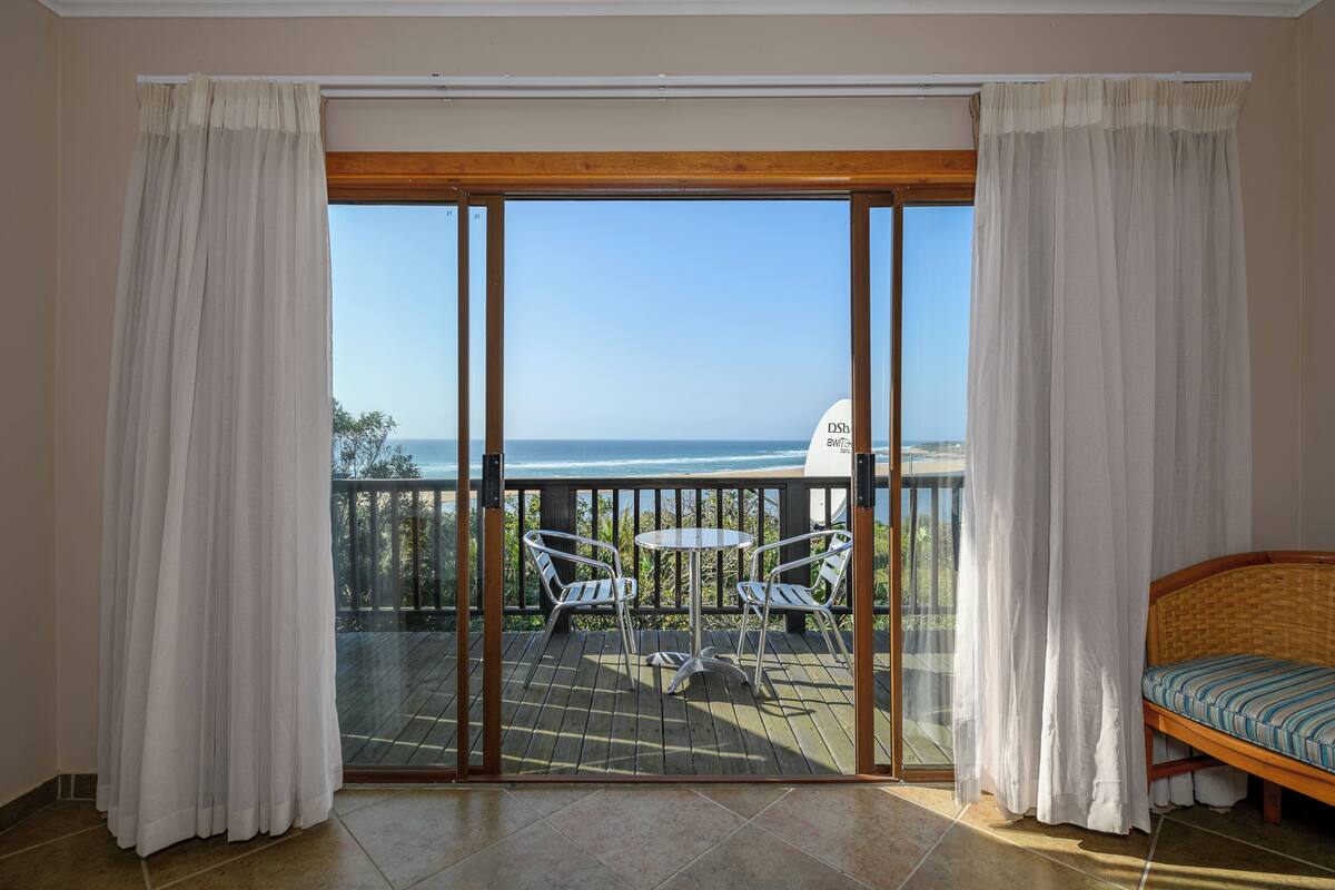 Una habitación con un gran ventanal corredera que da a un balcón con vistas al océano. Hay una pequeña mesa redonda con dos sillas en el balcón.