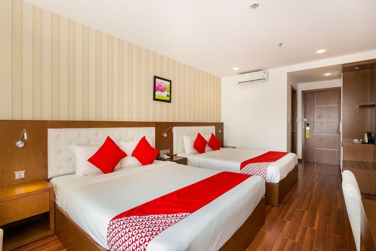A hotel room with two beds, a wooden headboard, a red and white bedspread, a nightstand, a wall-mounted air conditioner, and a framed picture.