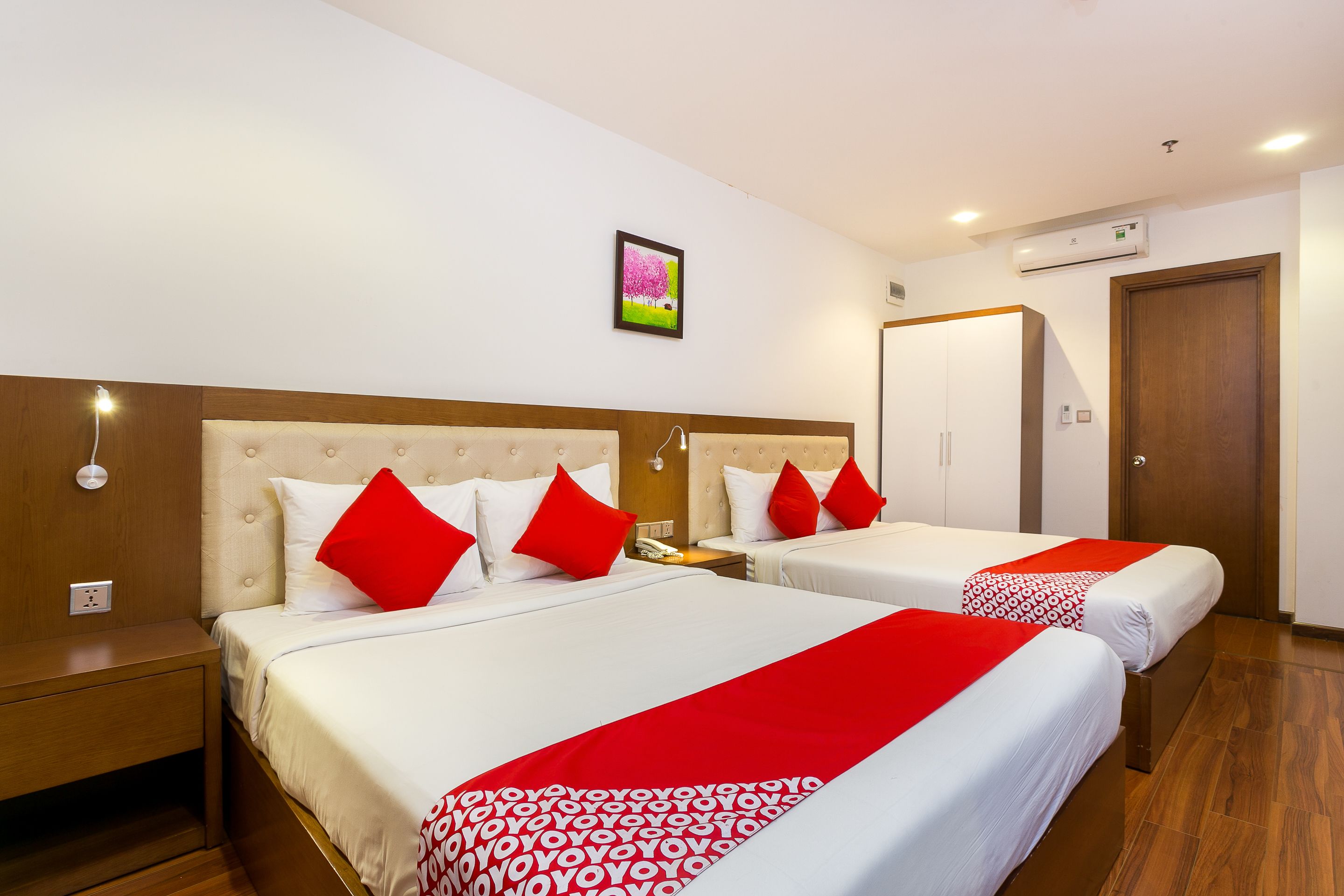 A hotel room with two beds, each with white bedding and red pillows. There is a painting on the wall, a bedside table with a lamp, and an air conditioning unit above the door.