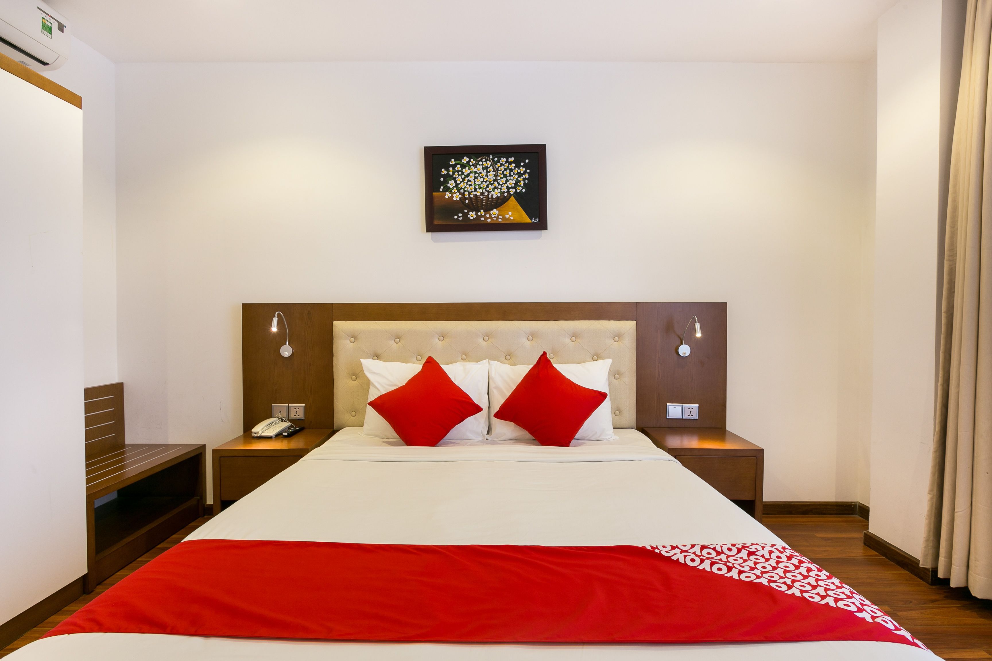 Habitación de hotel con una cama grande, dos almohadas rojas, una colcha blanca, un plaid rojo, una mesita de noche con un teléfono y una obra de arte colgada en la pared.