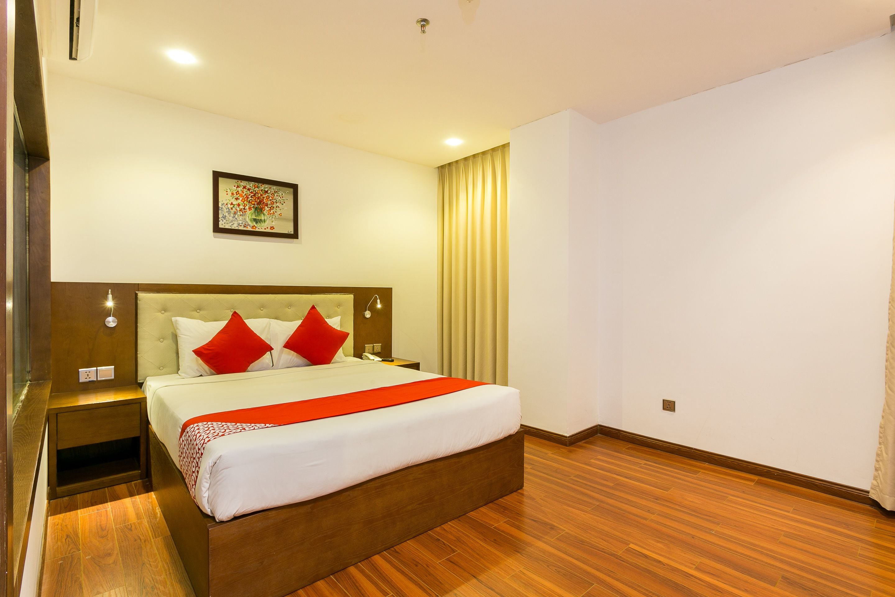 A hotel room with a large bed, wooden flooring, a nightstand, a bedside table, and a wall-mounted light fixture.