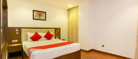 A hotel room with a large bed, wooden flooring, a nightstand, a bedside table, and a wall-mounted light fixture.
