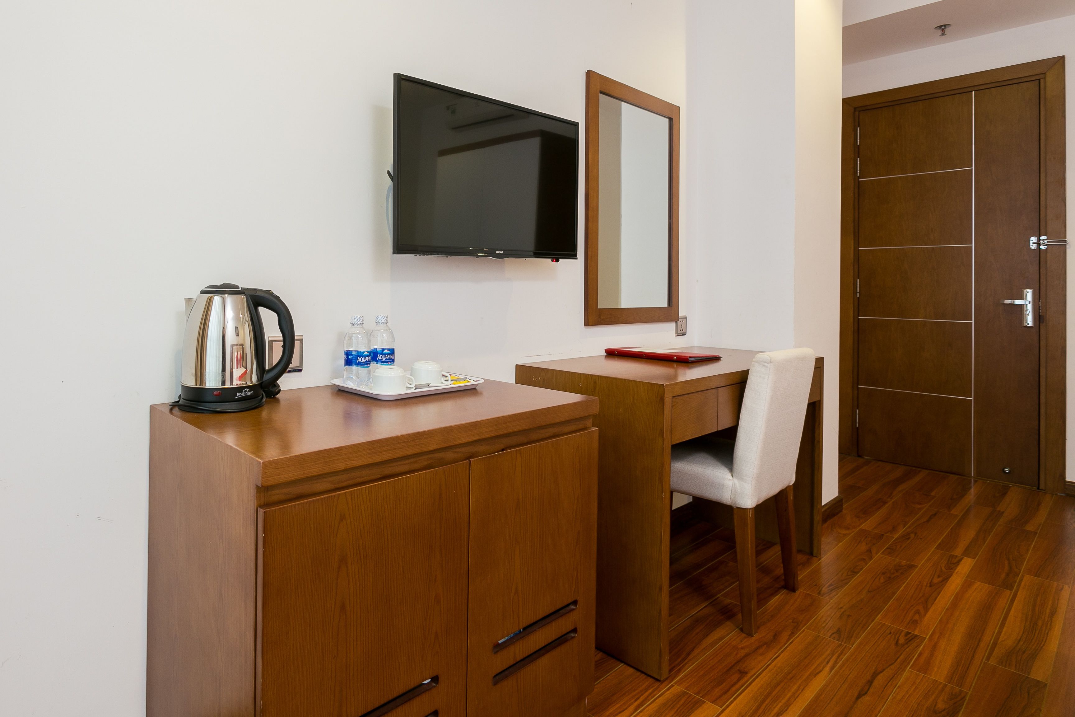 A hotel room with a wooden desk, a television mounted on the wall, a mirror, and a door.