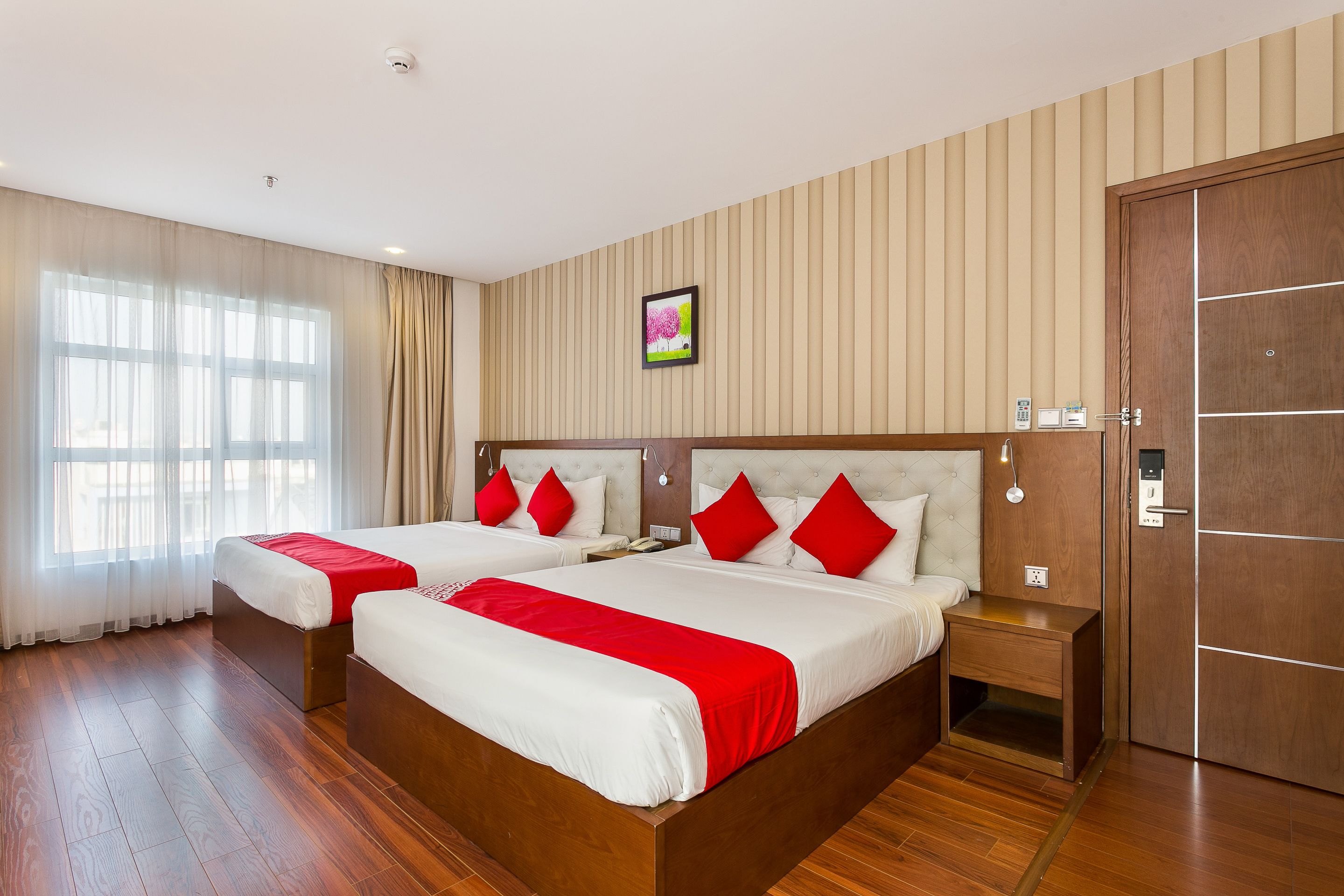 Camera d'albergo con due letti, pavimento in legno, un'ampia finestra con tende, un quadro appeso al muro e una porta con maniglia.