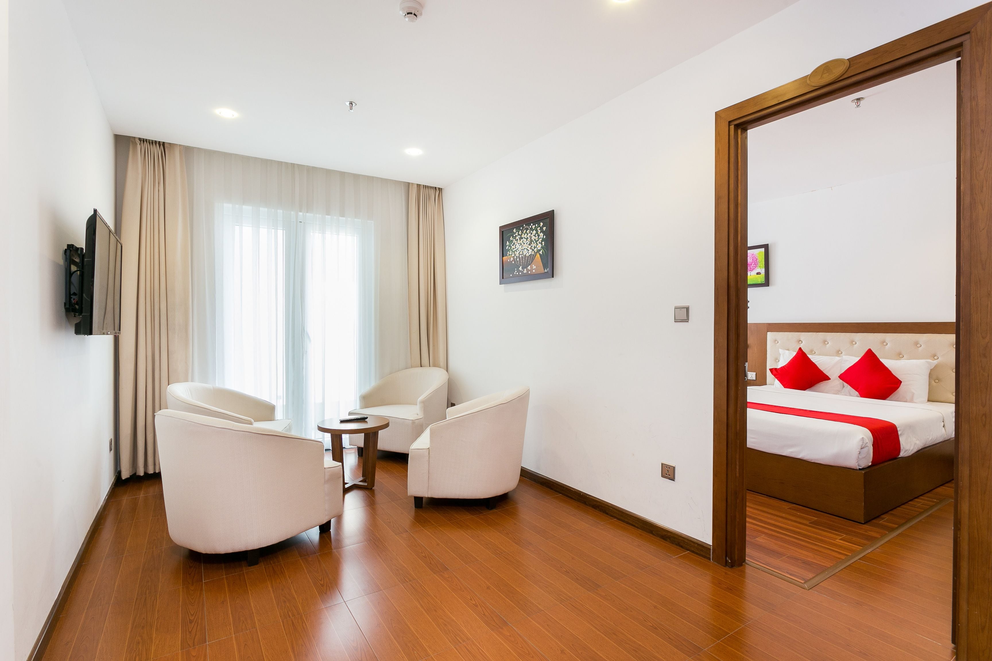 A hotel room with a wooden floor, a bed with red pillows, a TV on the wall, and two white armchairs.