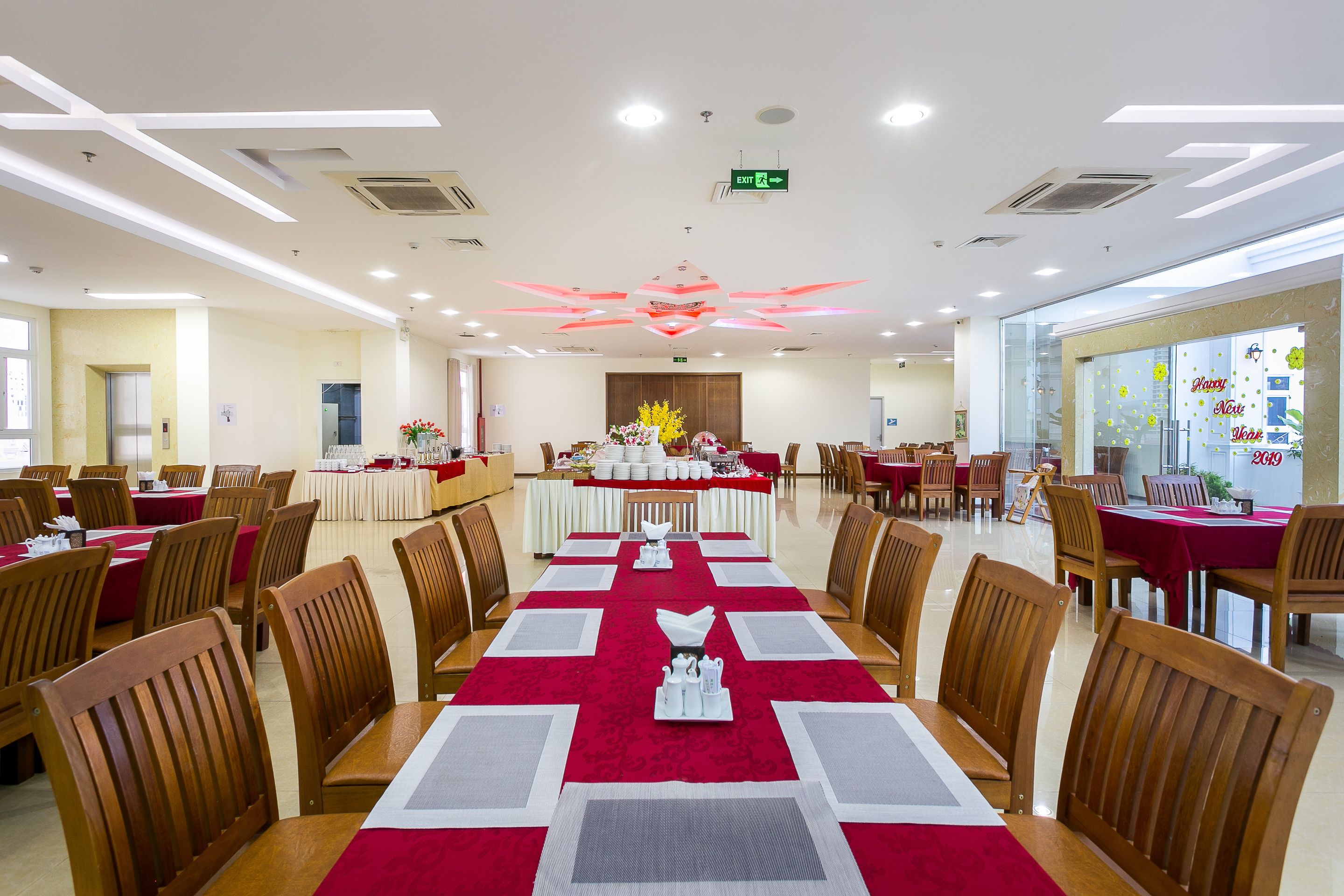 Une salle de banquet spacieuse, avec de nombreuses tables dressées pour le repas, un arrangement floral central et un design de plafond moderne.