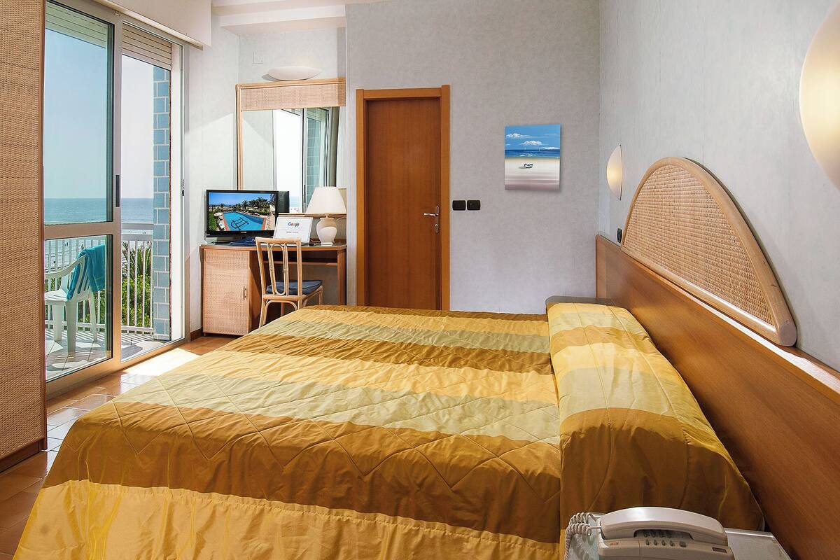 Una camera d'albergo con un letto, una scrivania con un computer, una sedia, un telefono e vista sul mare.