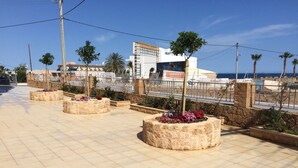 A paved walkway with stone planters containing flowers, a stone wall, and a view of a beach and buildings in the background. - Artemis Bay by Estia (Hersonissos)