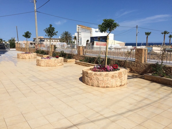 A paved walkway with stone planters containing flowers, a stone wall, and a view of a beach and buildings in the background.
