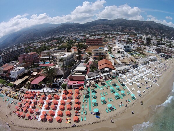 A coastal resort with multiple buildings, a beach with sun loungers and umbrellas, and a mountainous backdrop.