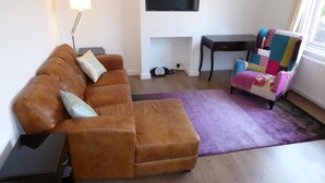 A living room with a brown sofa, a small side table, a colorful armchair, a flat-screen TV mounted on the wall, and a purple rug.