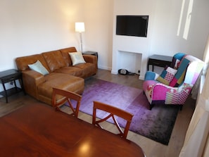 A living room with a brown leather sofa, a wooden dining table, a colorful armchair, a purple rug, and a flat-screen TV mounted on the wall. - Sunninghill Apartment (Ascot)