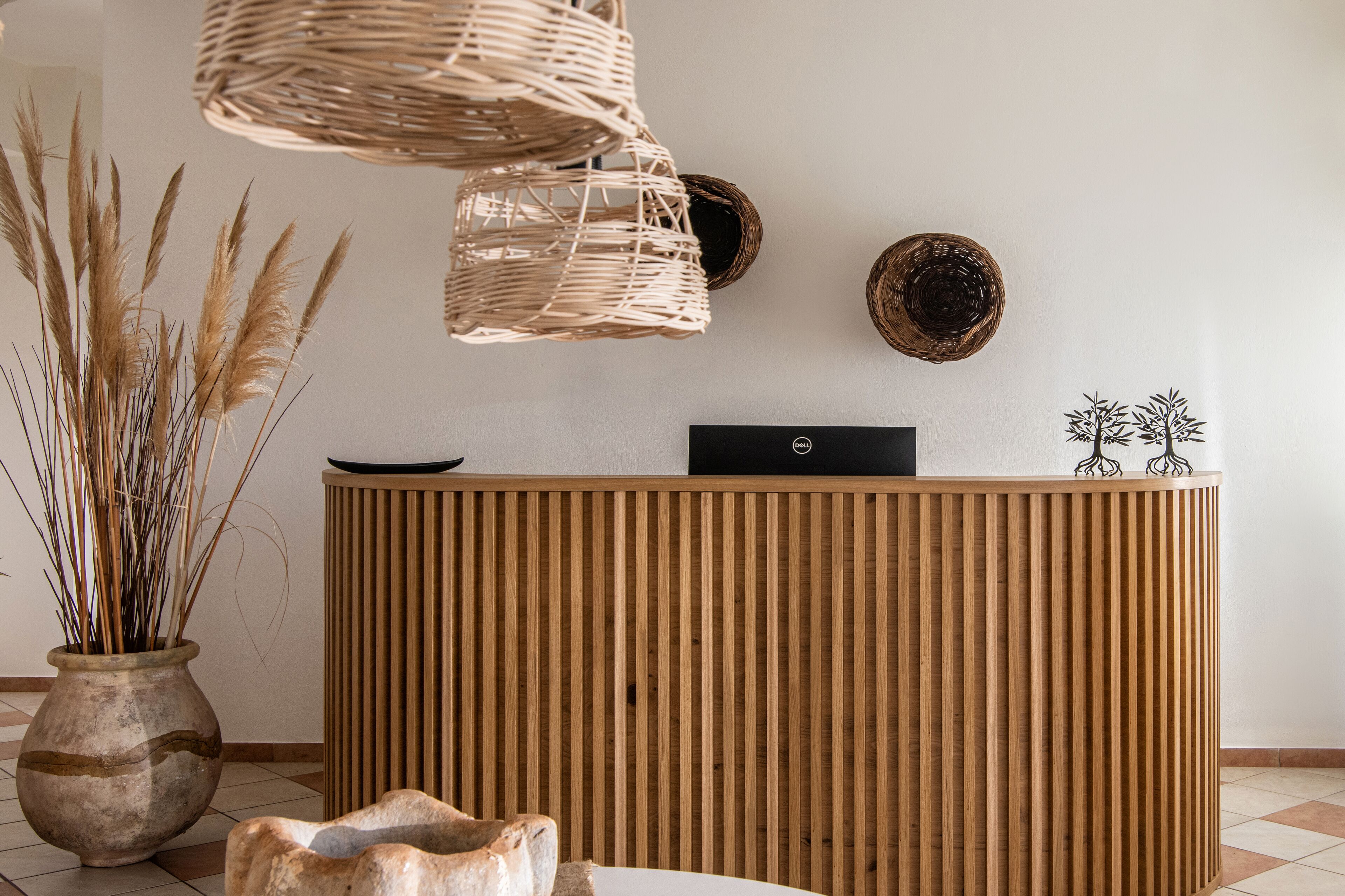 A reception desk with a wooden finish, a woven light fixture, a vase with dried plants, and a decorative plate on the wall.