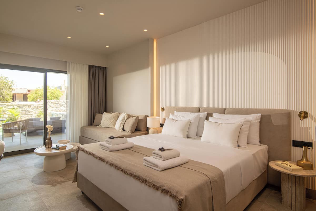 A modern hotel room with a large bed, a seating area with a table, and a view through the glass door.