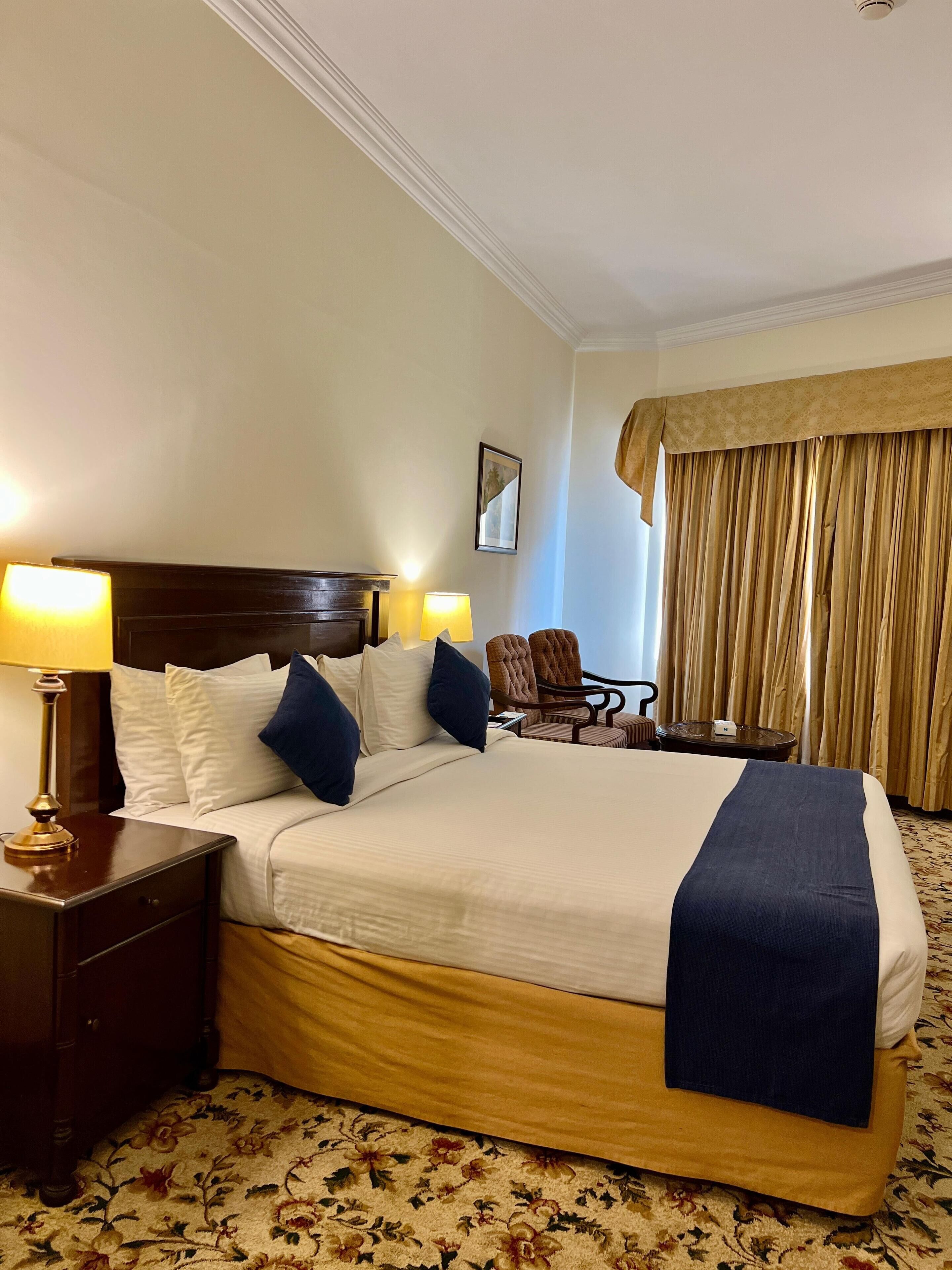 Een hotelkamer met een groot bed, twee nachtkastjes met lampen, een stoel, een kleine tafel en een raam met gordijnen.