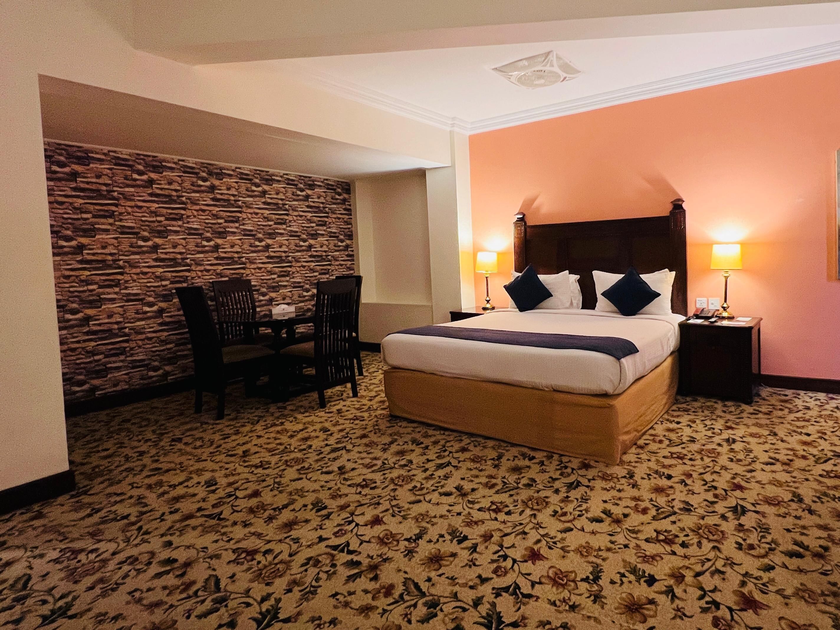 A hotel room with a large bed, two bedside lamps, a stone wall, a small table with chairs, and a floral carpet.