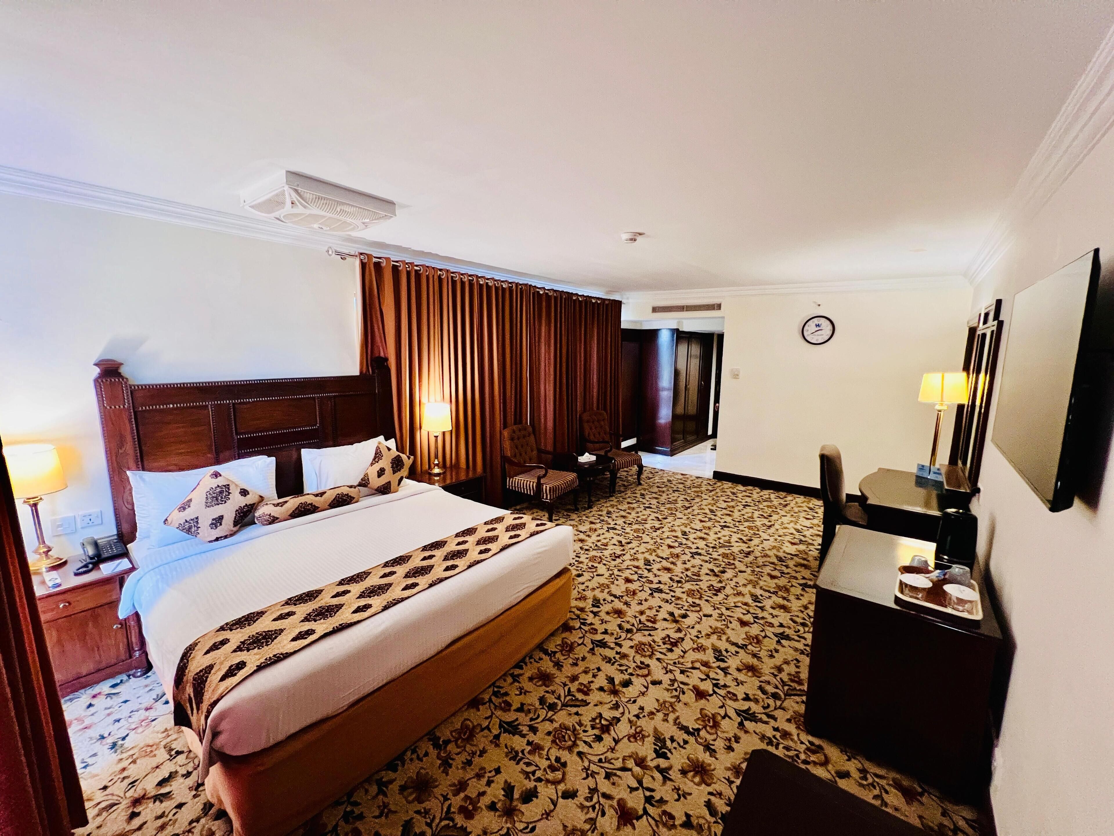 A hotel room with a large bed, a desk, a chair, a mirror, and a clock on the wall.