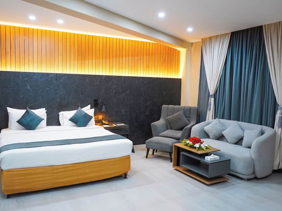 A modern hotel room with a large bed, a sofa, a small table with flowers, and a headboard with built-in lighting.