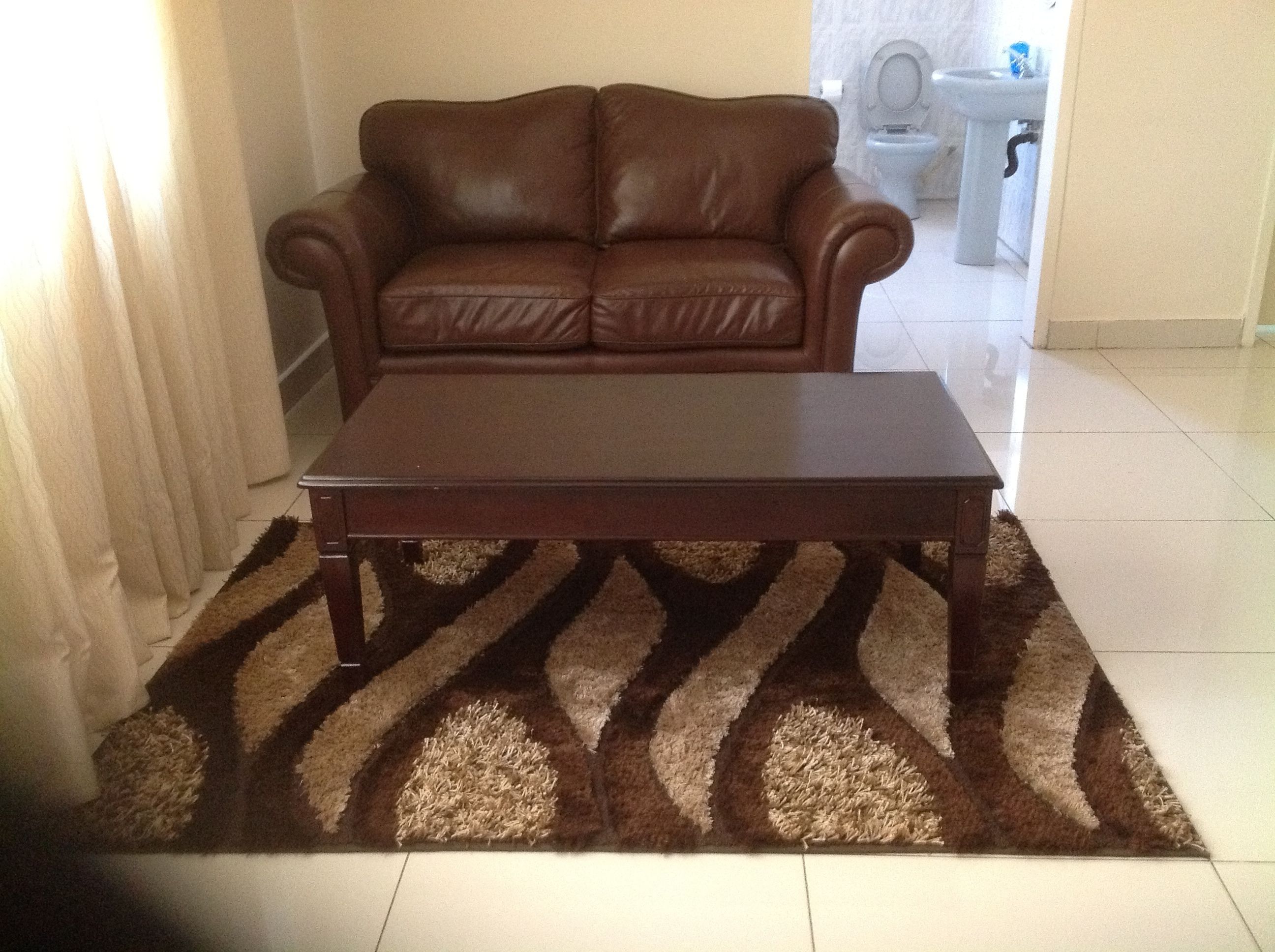 A brown leather sofa and a wooden coffee table on a patterned rug in a room with a white tiled floor and a bathroom in the background.