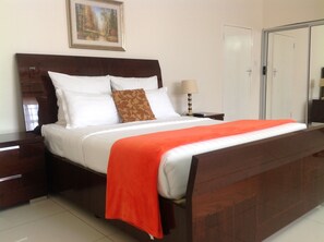 A neatly made bed with white and orange bedding, a wooden headboard, and a nightstand with a lamp. - Cypress Executive lodge (Livingstone)