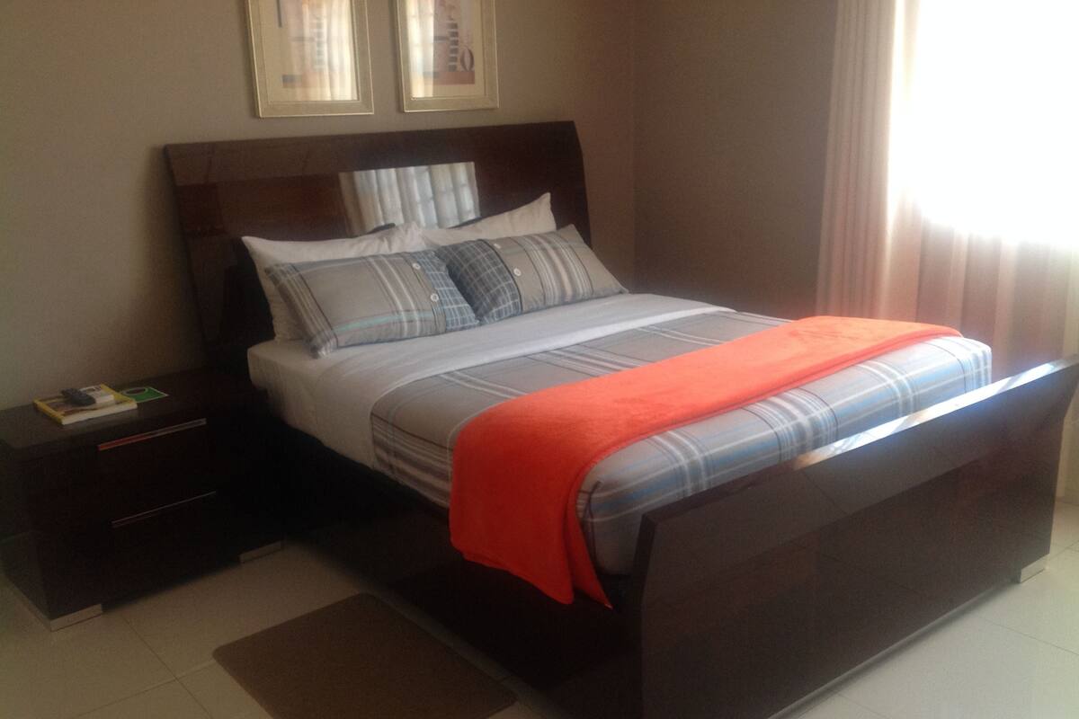 A neatly made bed with a plaid bedspread and pillows, a wooden nightstand with a book and a remote control, and a brown rug on the floor.