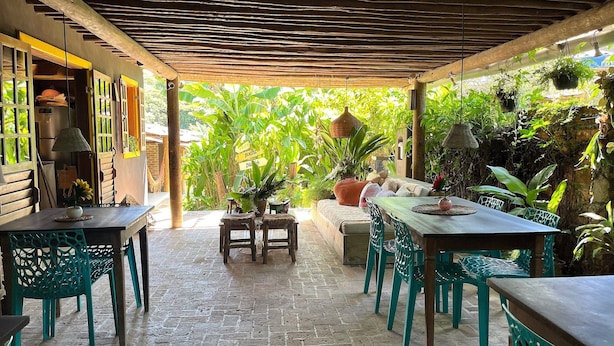 An outdoor patio with a rustic wooden ceiling, green chairs, a table with a vase of sunflowers, and lush greenery in the background. - Biribandos Rooms by Trancoso House (Porto Seguro)