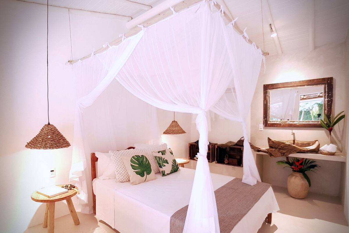 A bedroom with a canopy bed, a bedside table, a small stool, a mirror, and a potted plant.