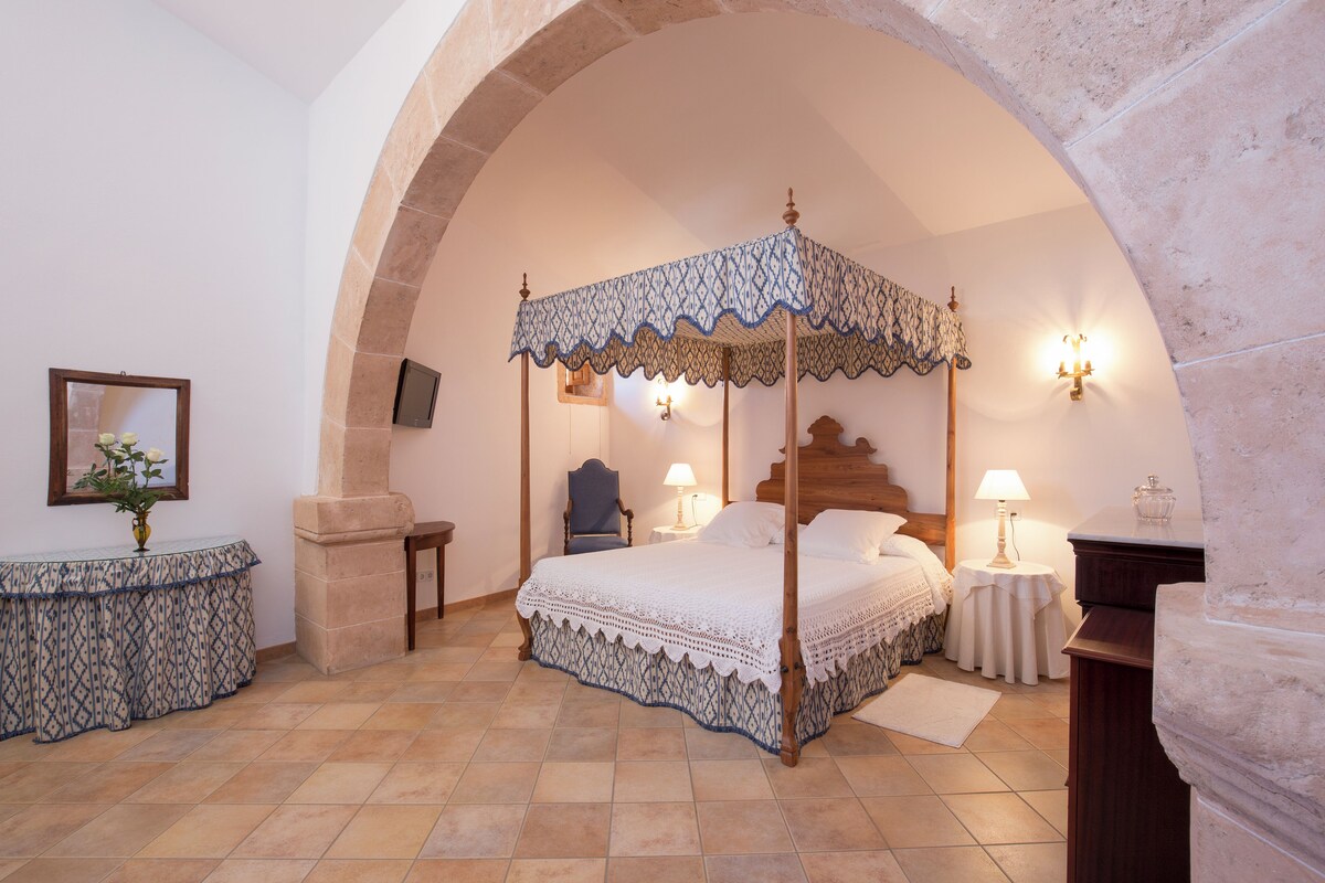 Un lit à baldaquin orné d’une voilette en dentelle, une arche en pierre, une table de chevet avec une lampe, et une petite table ronde également dotée d’une lampe.
