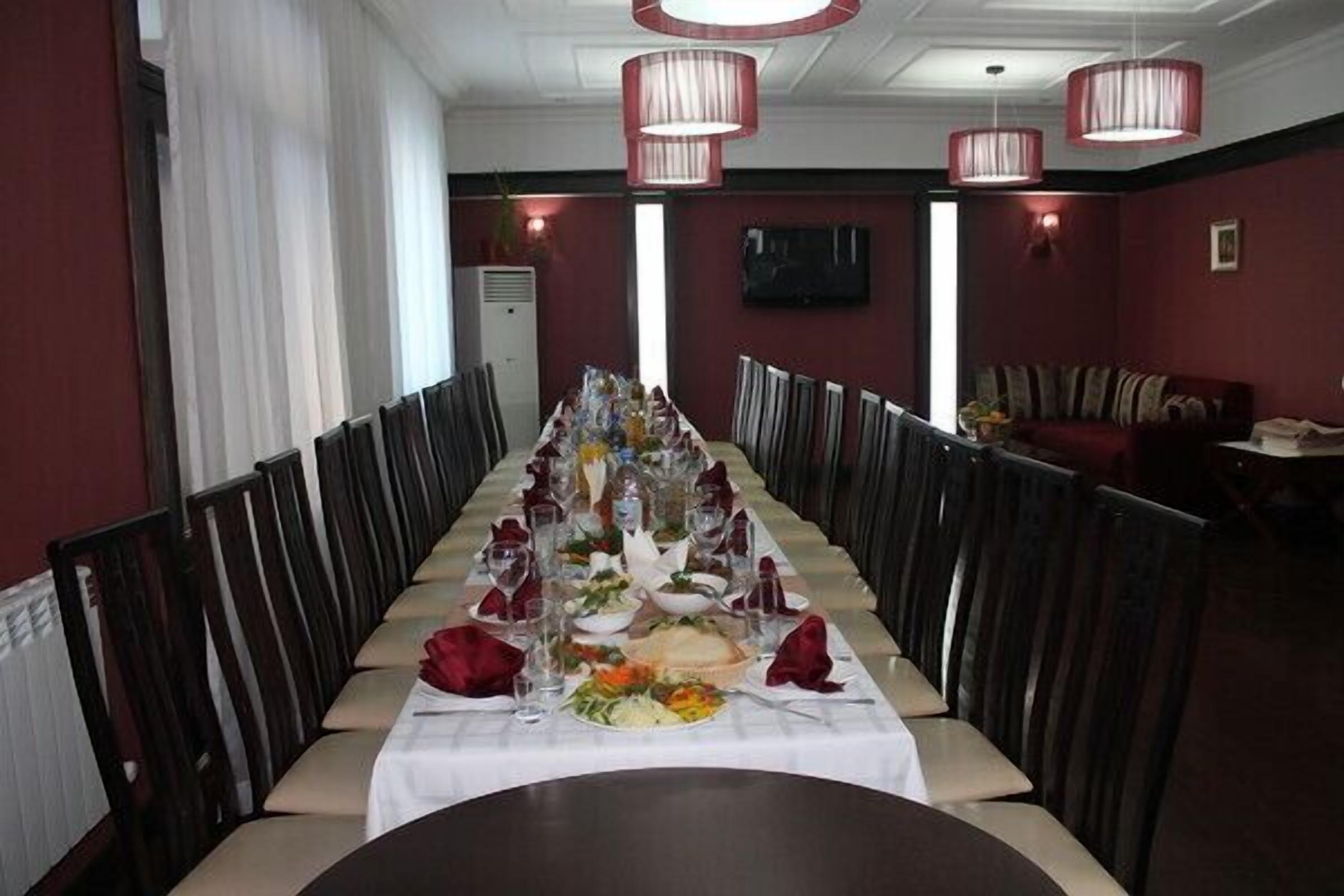 A long dining table set for a meal with chairs on both sides, a television on the wall, and a sofa in the background.