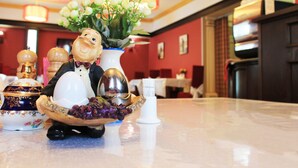 A dining table with a decorative ceramic figure, a vase with flowers, and a bowl of grapes. - Hotel San Marino (Astana)