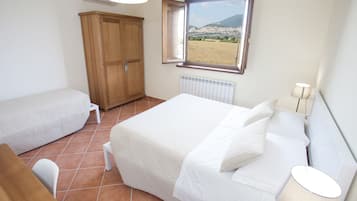A bedroom with a bed, a wooden wardrobe, a radiator, a window with a view of a city and mountains, and a table with a lamp.