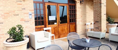 A brick building with a wooden door and a sign, outdoor seating with white chairs and tables, and potted plants.