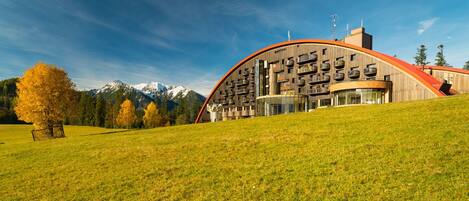 A modern building with a curved roof, large windows, and balconies, set against a mountainous backdrop.