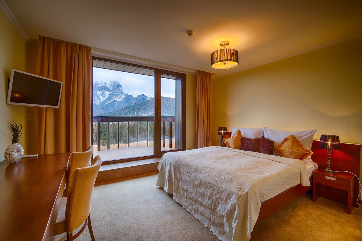 A hotel room with a large bed, a desk with a chair, a TV, and a view of mountains through a large window.