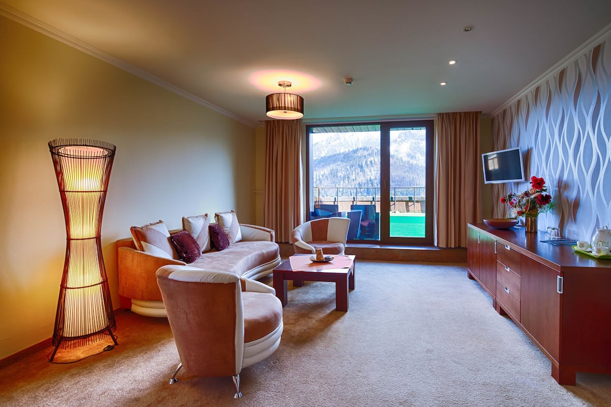 A hotel room with a sofa, armchairs, a coffee table, a TV, and a view of mountains through the window.