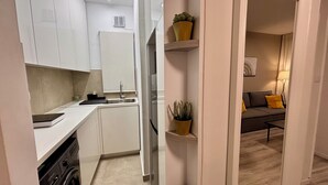 Una cocina moderna con armarios blancos, un fregadero y un lavavajillas. Hay dos plantas en macetas sobre estantes. Se ve un sofá y una lámpara en el área de estar contigua.
