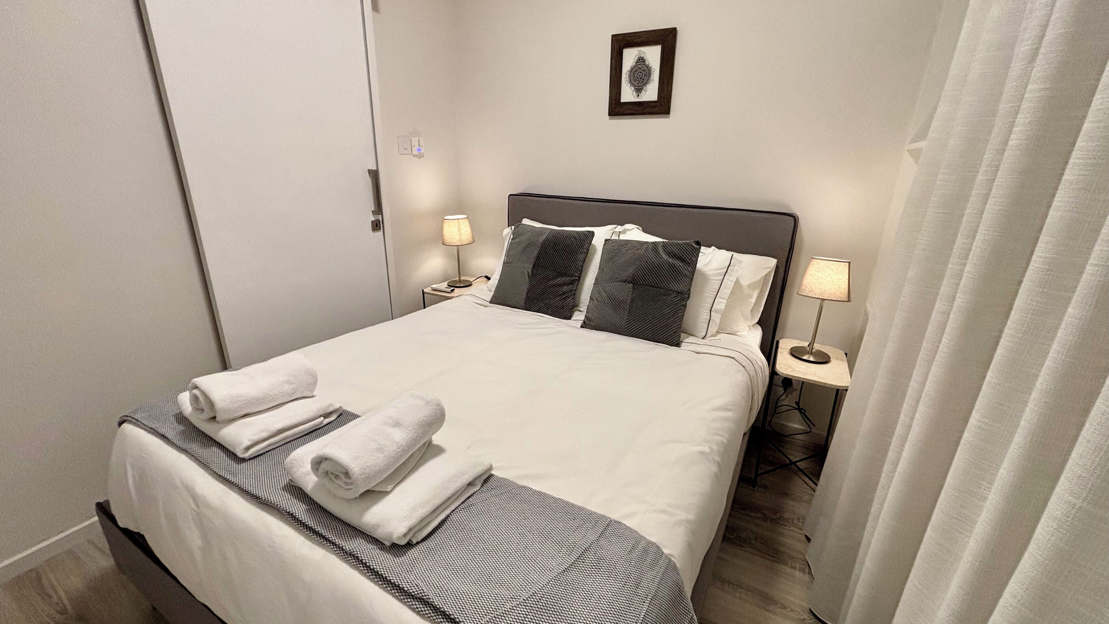 Un lit bien fait, avec du linge de lit blanc et des serviettes pliées, deux lampes de chevet et un tableau encadré au mur.