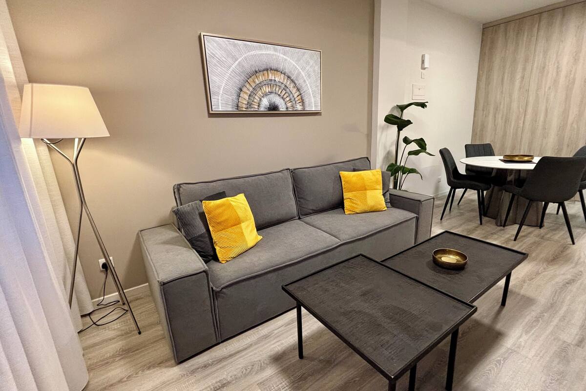 A modern living room with a grey sofa, two yellow cushions, a small coffee table, a floor lamp, a dining table with chairs, and a plant.