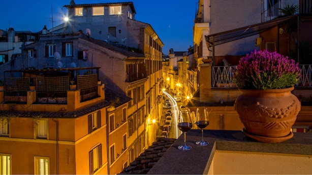 A nighttime view of a narrow street with buildings on either side, a potted plant with flowers, and two wine glasses on a ledge. - Palazzo Medusa (Rome)