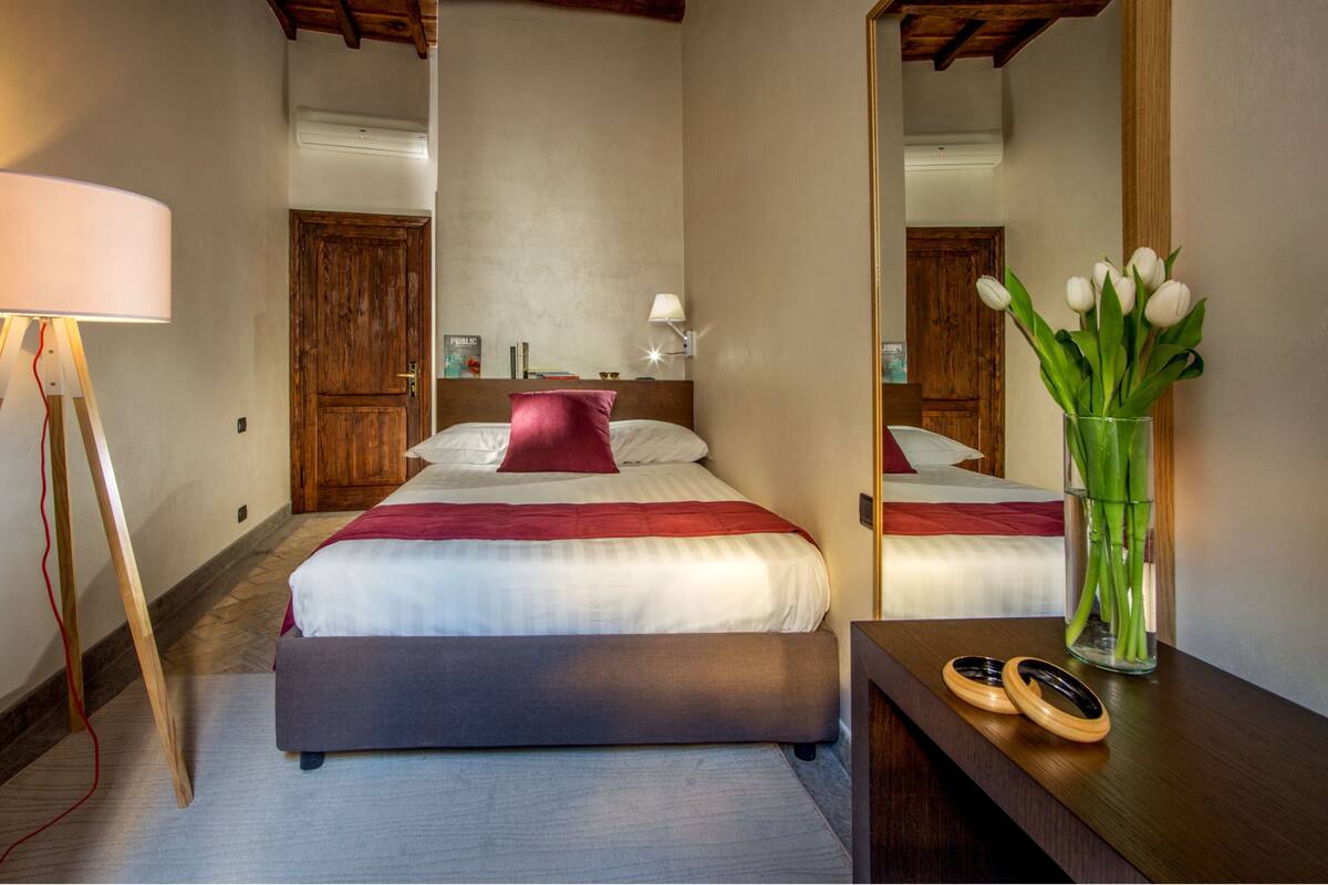 A hotel room with a bed, a mirror, a nightstand, a lamp, and a vase with flowers.
