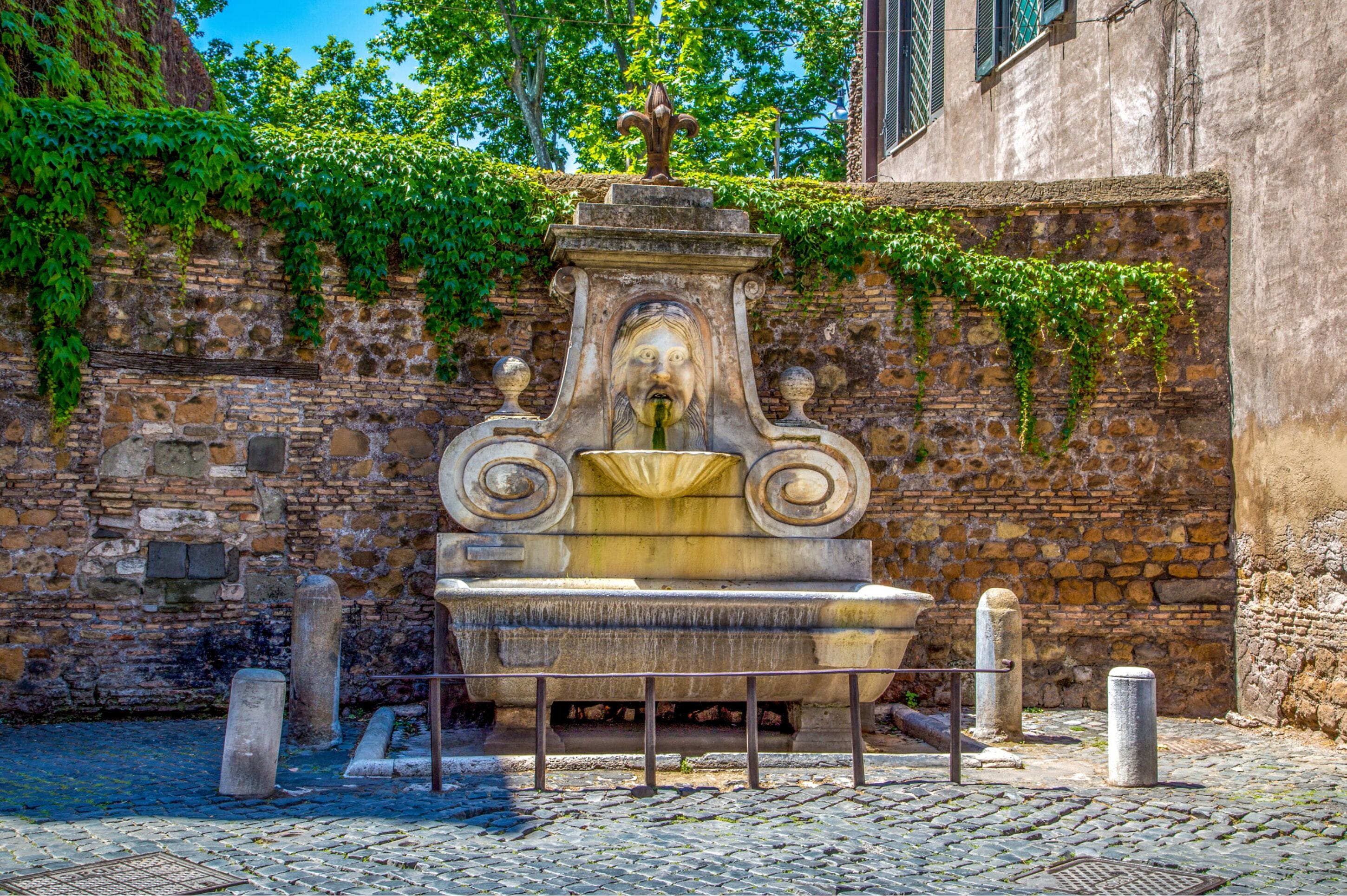 A stone fountain with a decorative top, surrounded by cobblestone pavement and a stone wall with ivy.