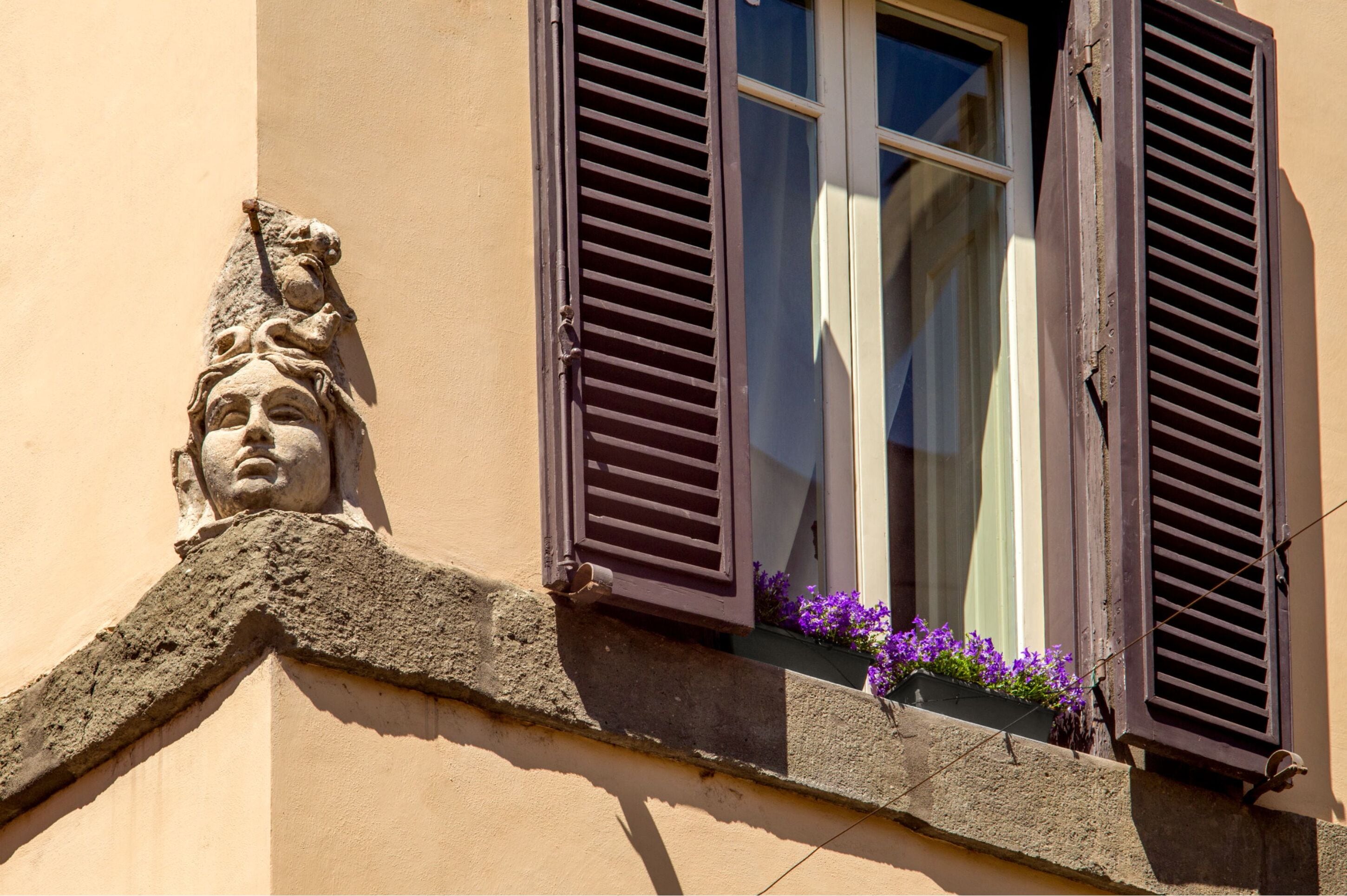 Un coin d’immeuble orné d’une sculpture en pierre, une fenêtre avec des volets et des fleurs violettes dans une jardinière.