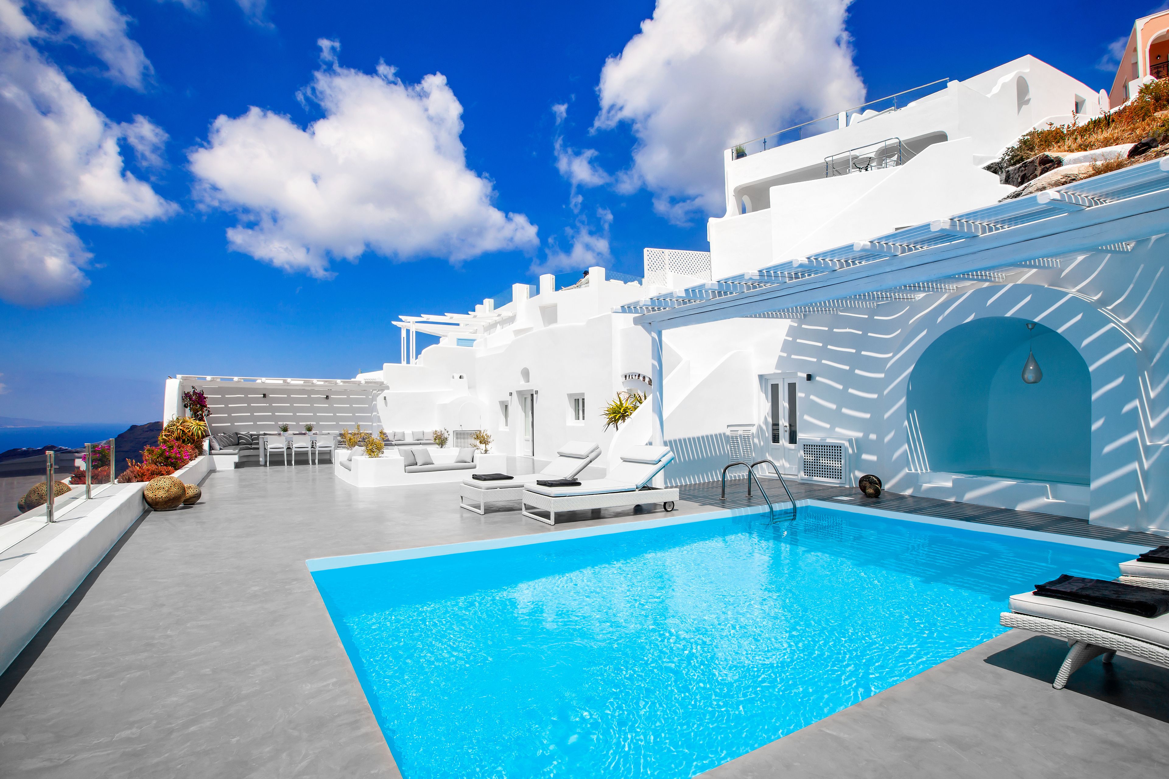 A modern pool area with white architecture, a clear blue sky, and a view of the sea.